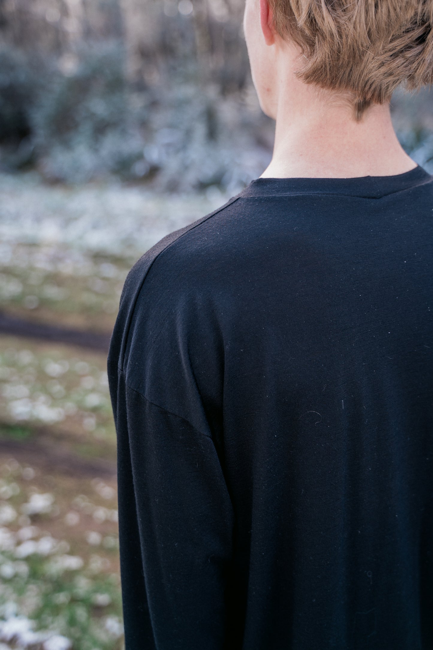 A man standing in a landscape lightly dusted with snow wearing a black shirt with his back to the camera showing shoulder detail.