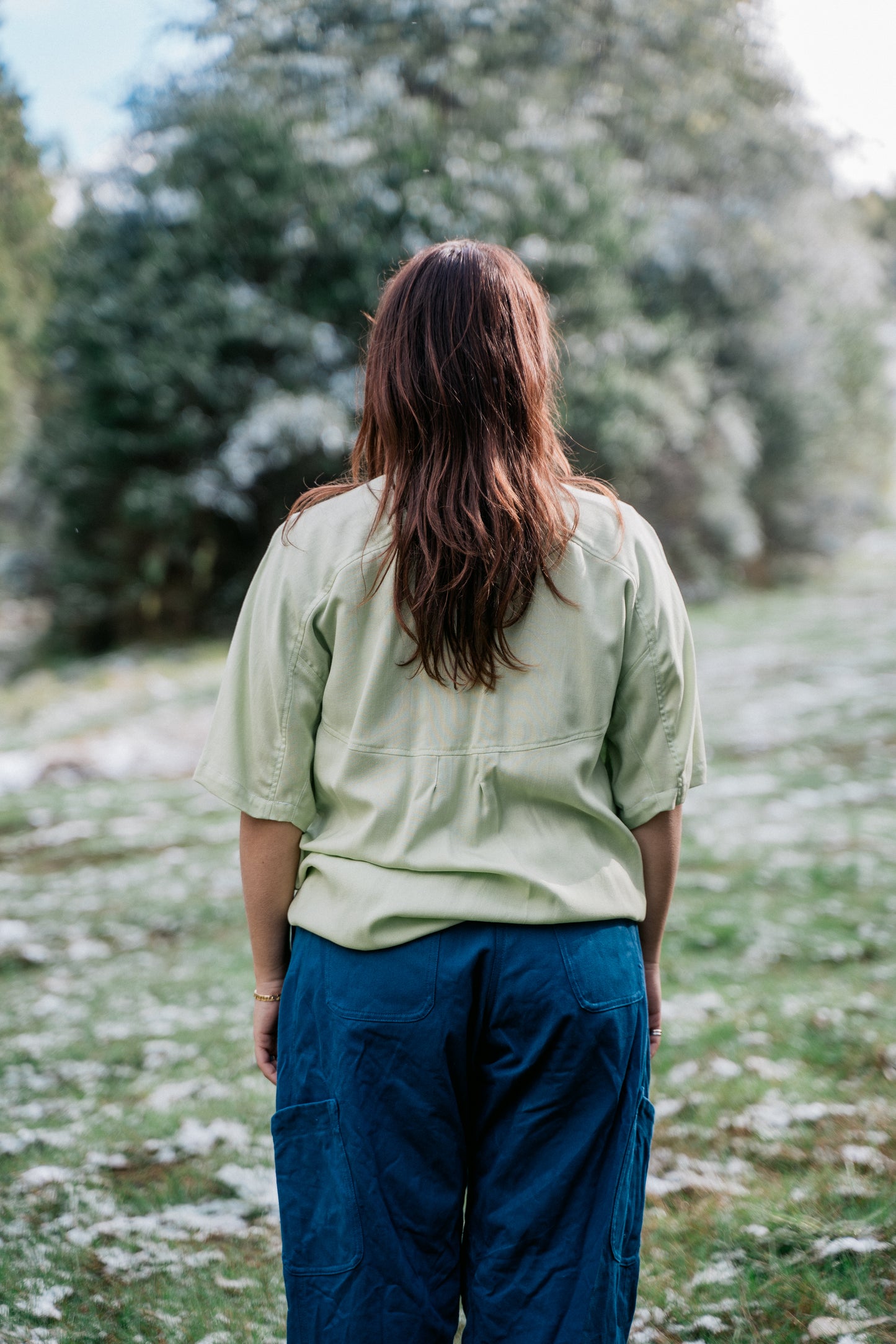 A woman wearing a light green collared shirt facing back to the camera in a grassy clearing covered in a light dusting of snow.