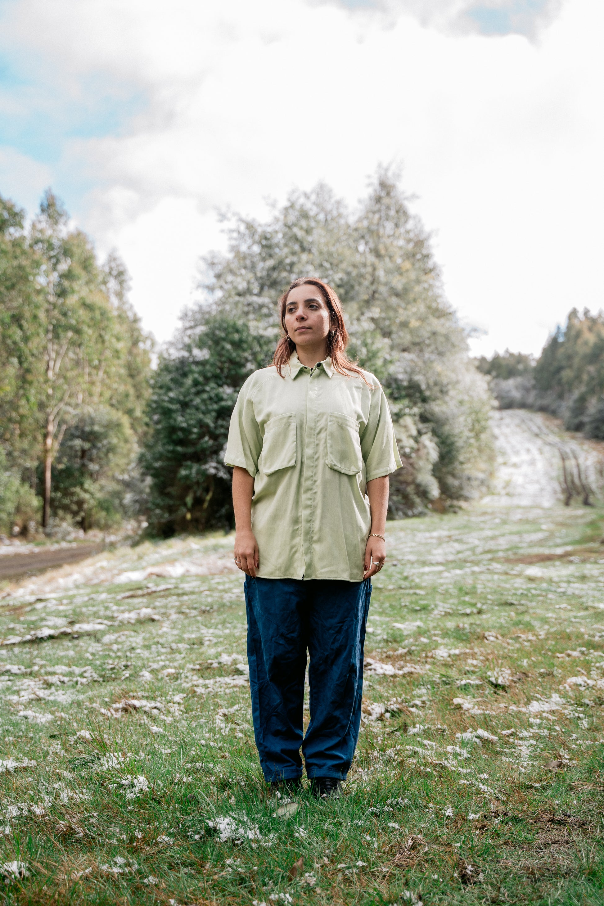 A woman wearing a light green collared shirt facing front to the camera in a grassy clearing covered in a light dusting of snow.