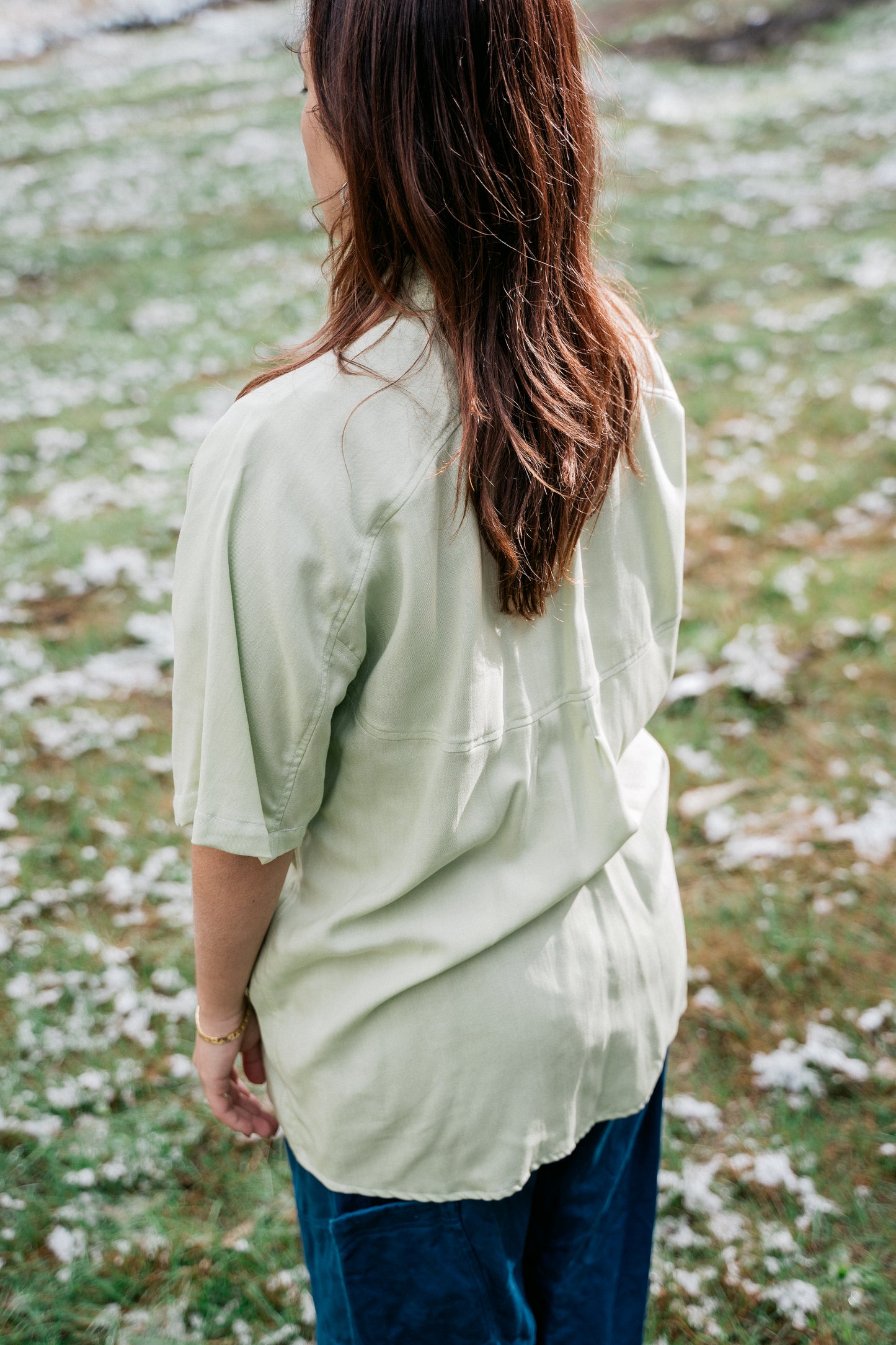 A woman wearing a light green collared shirt facing backwards from the camera in a grassy clearing covered in a light dusting of snow.