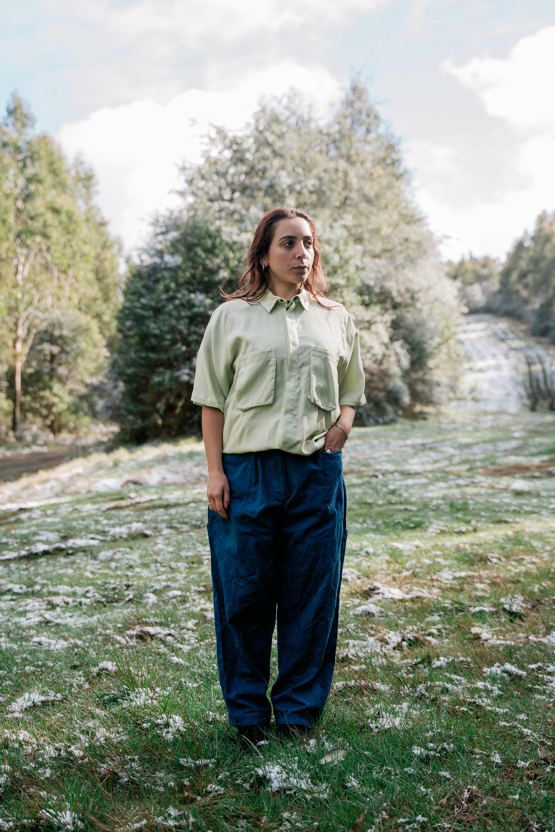 A woman wearing a light green collared shirt facing front to the camera in a grassy clearing covered in a light dusting of snow.