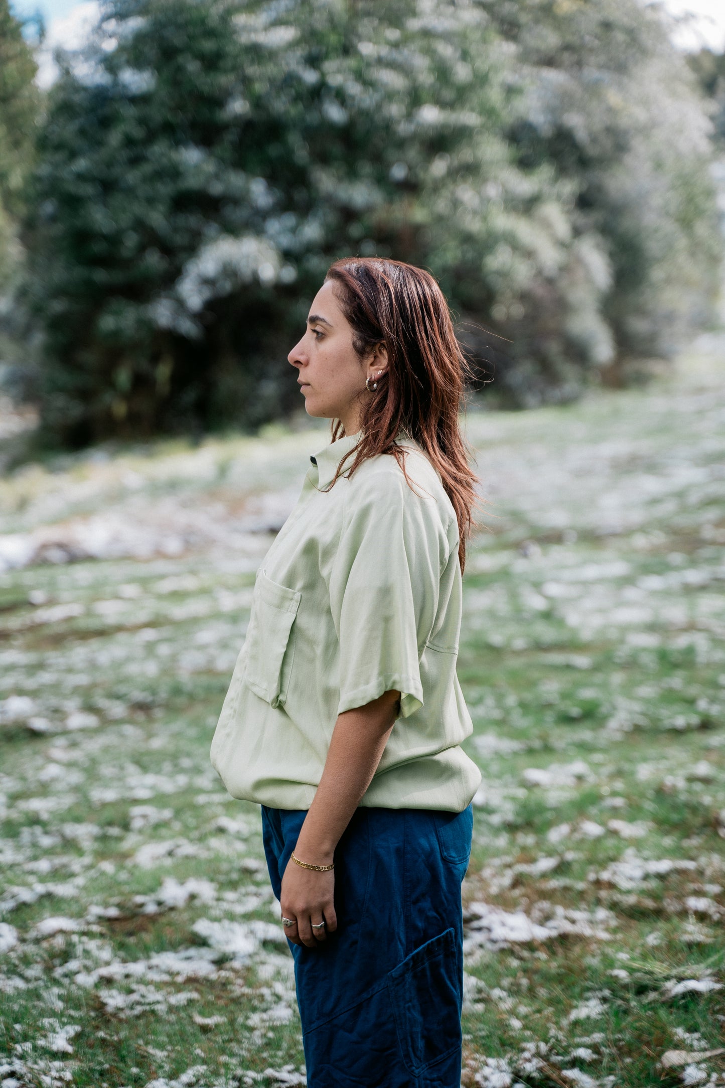 A woman wearing a light green collared shirt facing sideways from the camera in a grassy clearing covered in a light dusting of snow.