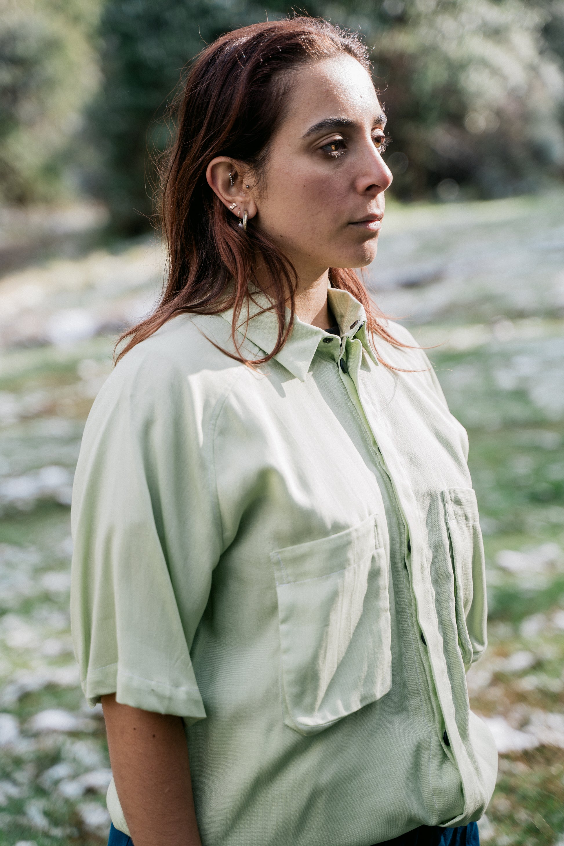 A woman wearing a light green collared shirt facing sideways from the camera in a grassy clearing covered in a light dusting of snow.