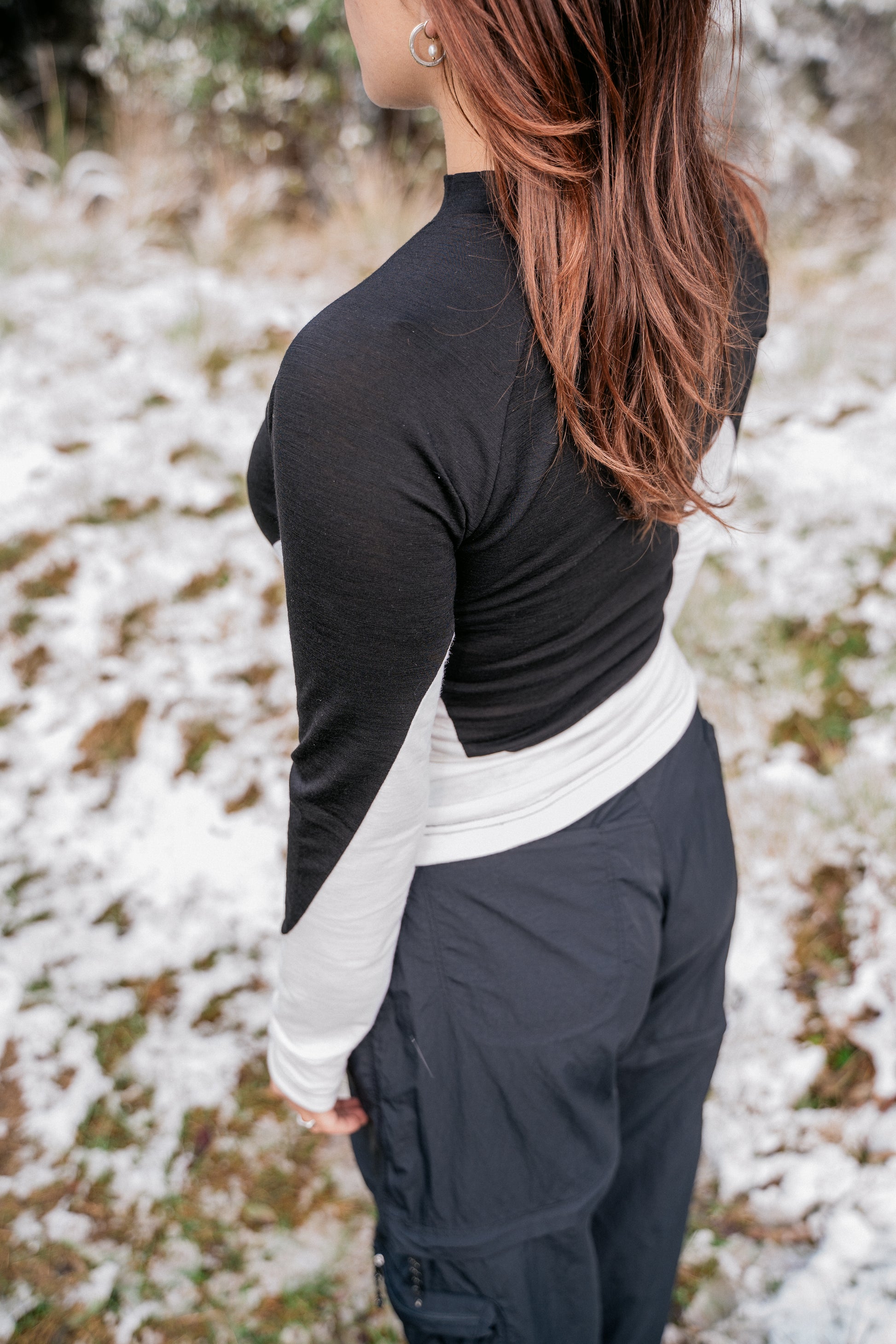 A woman wearing a half white-half black merino shirt in the snow.  Tha camera angle is looking down towards her shoulder.